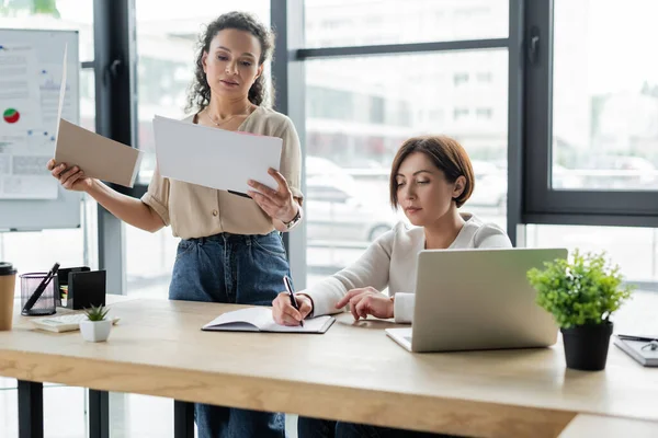 Afrikanisch-amerikanische Geschäftsfrau schaut sich Dokumente in der Nähe von Kollegen an, die am Arbeitsplatz in Notizbuch schreiben — Stockfoto