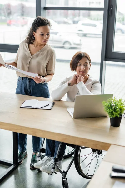 Afrikanisch-amerikanische Geschäftsfrau hält Papiere in der Hand und blickt auf Laptop neben Kollegin im Rollstuhl — Stockfoto