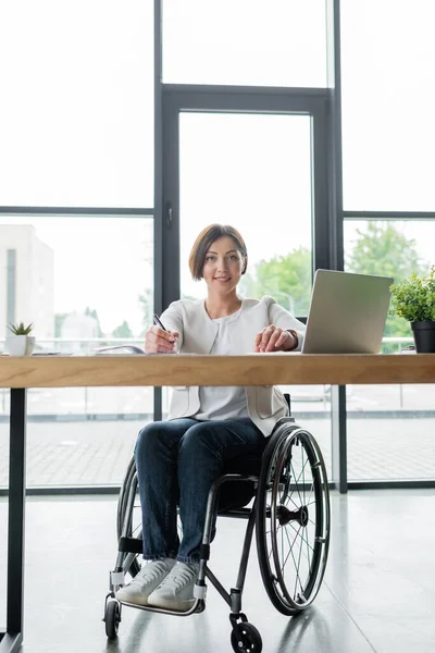 Erfolgreiche Geschäftsfrau im Rollstuhl lächelt in die Kamera, während sie in der Nähe ihres Laptops im Büro arbeitet — Stockfoto