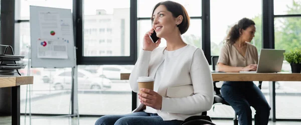 Mujer feliz con discapacidad sosteniendo café para ir mientras habla en el teléfono celular cerca de un colega afroamericano, pancarta - foto de stock