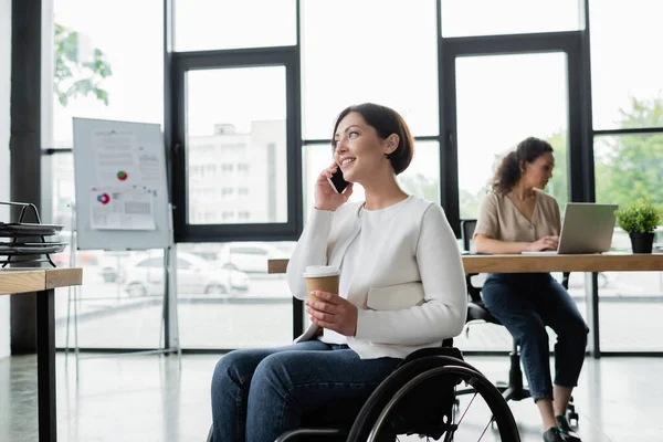 Улыбающаяся деловая женщина в инвалидной коляске держит кофе, чтобы пойти и поговорить по мобильному телефону рядом с размытым африканским американским коллегой — стоковое фото