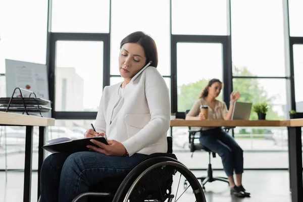 Femme d'affaires en fauteuil roulant écrit dans un carnet tout en parlant sur un téléphone portable dans le bureau — Photo de stock