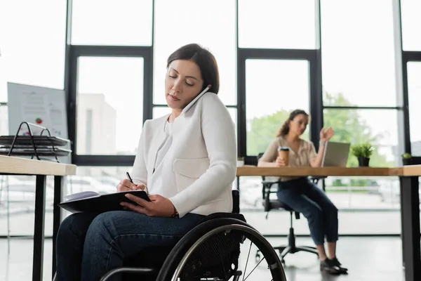 Empresaria en silla de ruedas escribiendo en cuaderno mientras habla por celular en la oficina - foto de stock