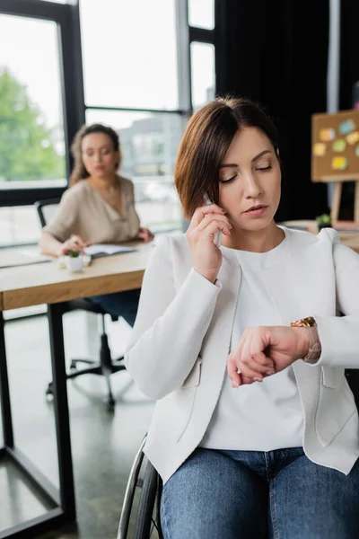 Mujer de negocios con discapacidad mirando reloj de pulsera durante la conversación telefónica cerca borrosa colega afroamericano — Stock Photo