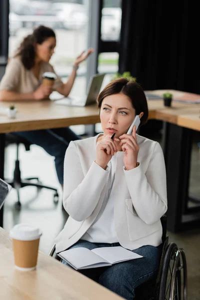 Femme d'affaires en fauteuil roulant tenant un stylo et parlant sur un téléphone mobile près d'un ordinateur portable vide dans le bureau — Photo de stock