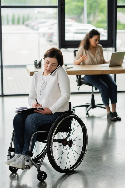 Businesswoman in wheelchair talking on mobile phone and writing in notebook near blurred african american colleague — Stock Photo