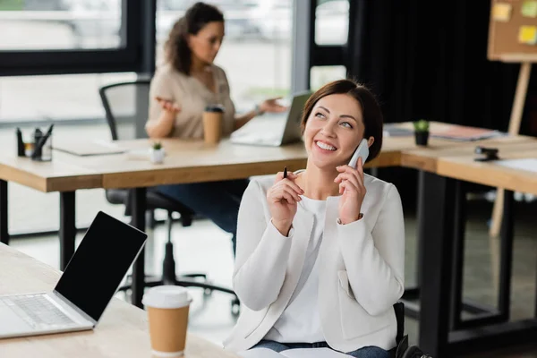 Mujer alegre con discapacidad hablando en teléfono inteligente cerca de la computadora portátil con pantalla en blanco y difuminado colega afroamericano - foto de stock