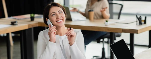 Allegra donna d'affari che parla su smartphone vicino a laptop con schermo bianco in ufficio, banner — Foto stock