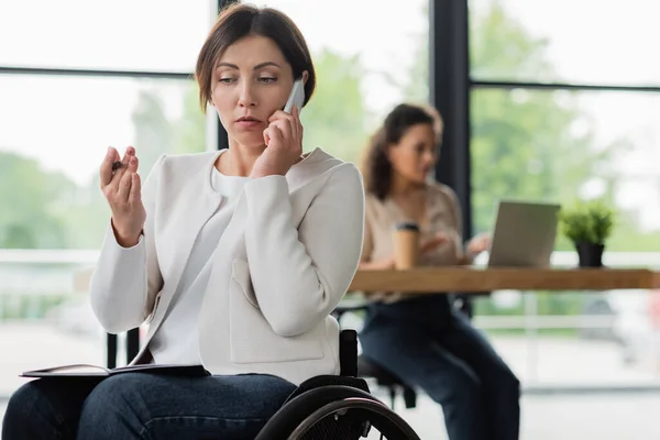 Businesswoman in wheelchair talking on mobile phone near african american woman working on blurred background — Stock Photo