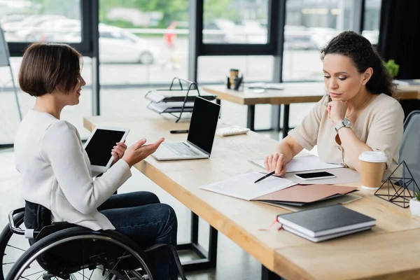 Lächelnde Geschäftsfrau im Rollstuhl im Gespräch mit nachdenklichen afrikanisch-amerikanischen Kollegen, die mit Dokumenten arbeiten — Stockfoto