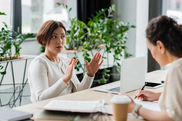 Businesswoman gesturing near laptop while talking to african american colleague writing on blurred foreground — Stock Photo