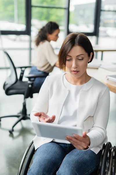 Donna d'affari sulla sedia a rotelle che punta al tablet digitale mentre lavora vicino a un collega afroamericano offuscato — Foto stock