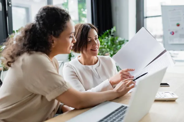 Mujeres de negocios interracial apuntando al documento mientras trabajan juntas en el cargo - foto de stock