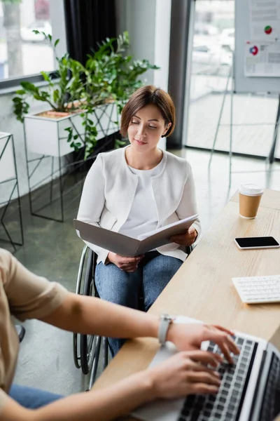 Donna d'affari positiva con disabilità guardando i documenti vicino collega afro-americano digitando sul computer portatile in primo piano sfocato — Foto stock