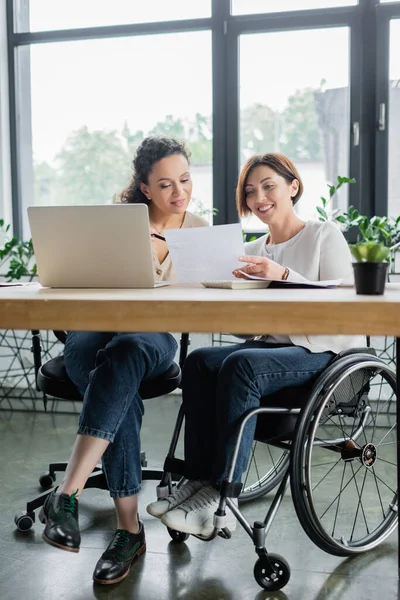 Smiling woman in wheelchair working with african american colleague in office — Stock Photo