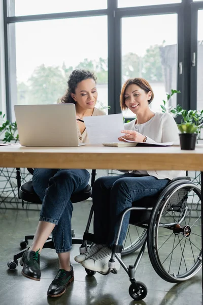 Mujer sonriente en silla de ruedas trabajando con un colega afroamericano en la oficina - foto de stock