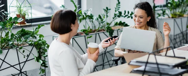 Mujer de negocios borrosa mostrando teléfono inteligente a alegre colega afroamericano en el lugar de trabajo en la oficina, pancarta - foto de stock