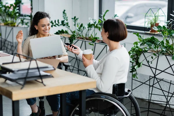 Businesswoman in wheelchair showing smartphone to smiling african american colleague at workplace — Stock Photo