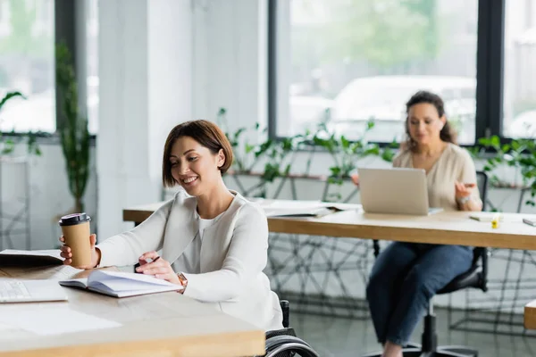 Happy businesswoman with disability holding paper cup while working near blurred african american colleague — Stock Photo