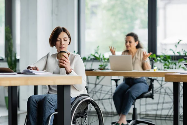 Donna d'affari in sedia a rotelle bere caffè per andare vicino collega afro-americano che lavora su sfondo sfocato — Foto stock