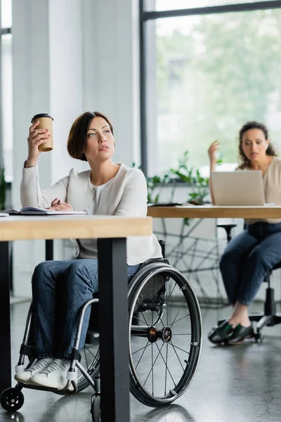 Pensativa empresária em cadeira de rodas segurando takeaway bebida perto de colega afro-americano borrado no escritório — Fotografia de Stock