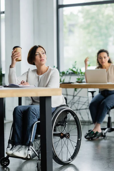 Femme d'affaires réfléchie en fauteuil roulant tenant boisson à emporter près flou confrère afro-américain au bureau — Photo de stock