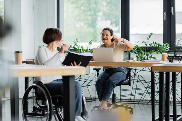 Businesswoman in wheelchair showing notebook to african american colleague pointing at laptop — Stock Photo