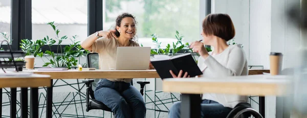 Feliz mujer de negocios africana apuntando a la computadora portátil cerca borrosa colega con discapacidad, pancarta — Stock Photo