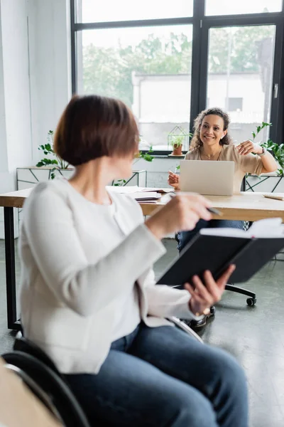 Blurred businesswoman with disability pointing at notebook near african american colleague working at laptop — Stock Photo
