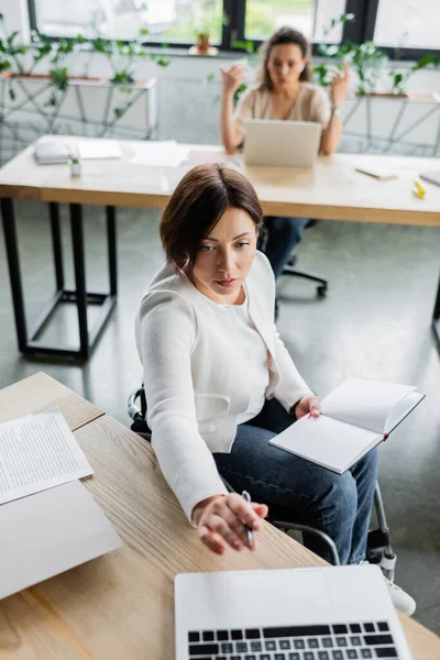 Businesswoman in wheelchair holding empty notebook while working near laptop and blurred african american colleague — Stock Photo