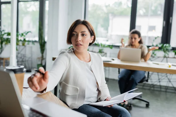 Businesswoman with physical disability pointing at blurred laptop while working near african american colleague — Stock Photo