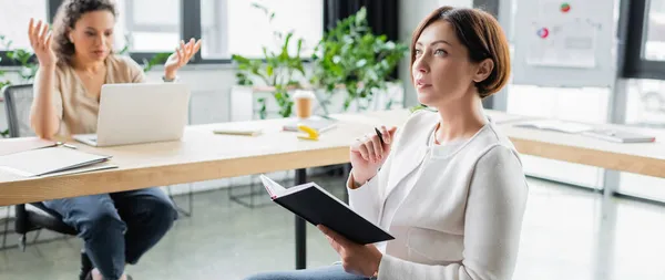 Pensativa mujer de negocios con portátil cerca borrosa colega afroamericano gesto mientras mira el ordenador portátil, pancarta - foto de stock