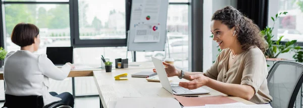 Mujer de negocios afroamericana positiva que tiene videollamada mientras trabaja cerca de colega en silla de ruedas, pancarta — Stock Photo