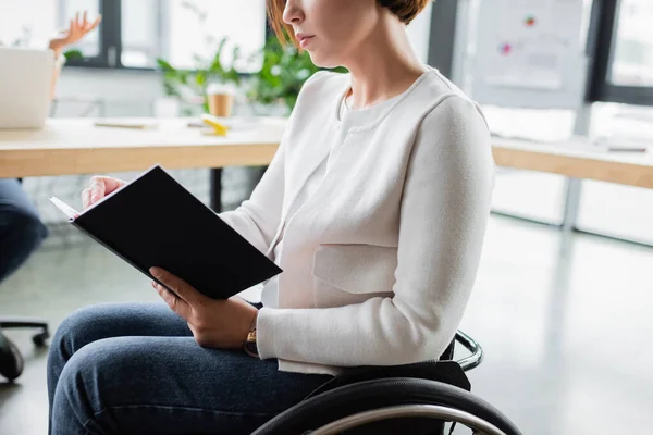 Обрізаний вид бізнес-леді в інвалідному візку тримає блокнот в офісі — стокове фото
