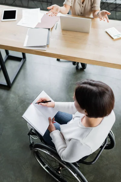 Vista aérea de una mujer de negocios en silla de ruedas escribiendo en un cuaderno vacío cerca de un colega haciendo gestos en el portátil - foto de stock