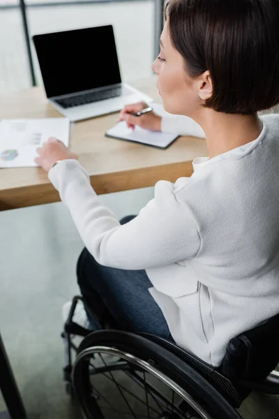 Donna d'affari con disabilità che lavora vicino a laptop sfocato con schermo bianco in ufficio — Foto stock