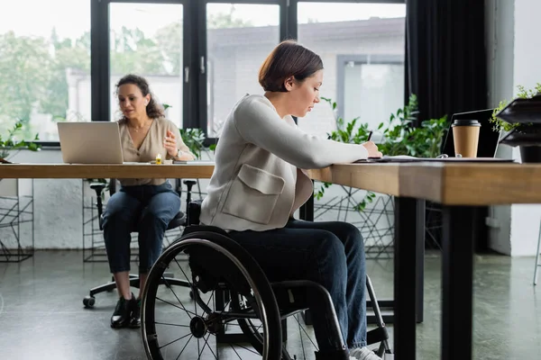 Mujer de negocios en silla de ruedas que trabaja en la oficina cerca de la computadora portátil y difuminado colega afroamericano - foto de stock