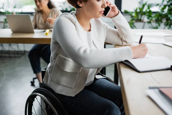 Businesswoman with disability talking on smartphone and writing in notebook in office — Stock Photo