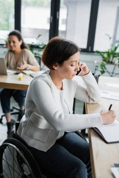 Femme d'affaires handicapée physique écrivant dans un carnet lors d'une conversation sur smartphone au bureau — Photo de stock