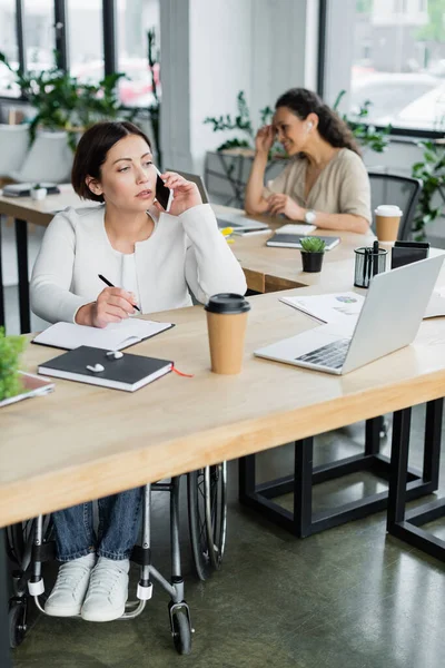 Businesswoman in wheelchair talking on mobile phone near laptop and blurred african american colleague — Stock Photo