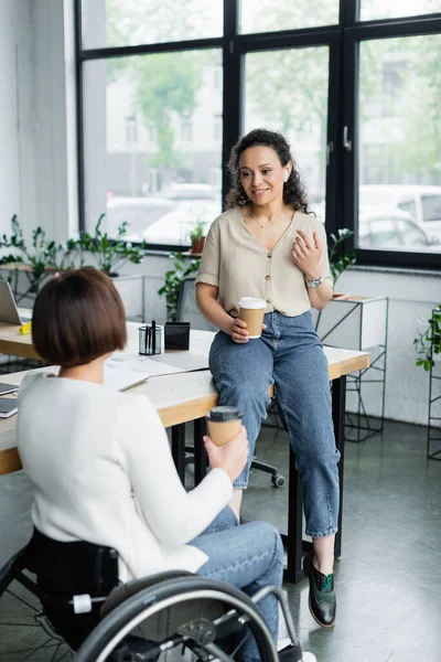 Una donna d'affari afroamericana sorridente seduta sulla scrivania mentre parla con un collega sulla sedia a rotelle durante una pausa caffè — Foto stock