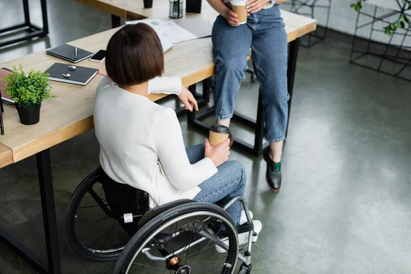 Donna d'affari in sedia a rotelle in possesso di tazza di carta vicino collega seduto sulla scrivania durante la pausa caffè — Foto stock