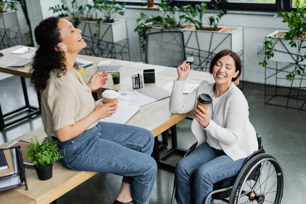 Emocionada mujer de negocios afroamericana y colega en silla de ruedas riendo durante el descanso de café - foto de stock