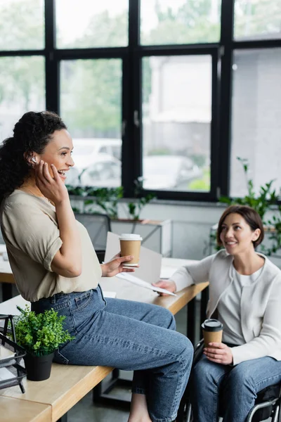 Allegra donna d'affari africana americana che aggiusta l'auricolare mentre si siede sulla scrivania vicino al collega in sedia a rotelle — Foto stock