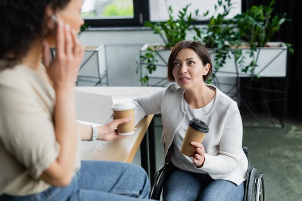 Mujer de negocios en silla de ruedas hablando con un borroso colega afroamericano sentado en el escritorio durante el descanso del café - foto de stock