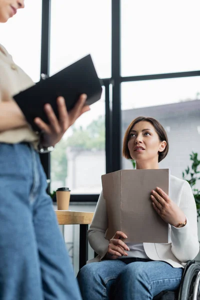 Businesswoman with disability holding documents and looking at african american colleague writing in notebook — Stock Photo