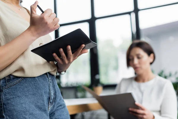 African american businesswoman holding notebook and pen near blurred colleague with paper folder — Stock Photo