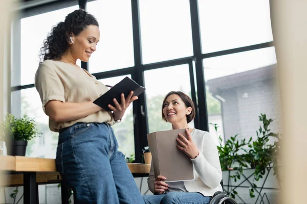 Feliz empresaria en silla de ruedas sosteniendo documentos cerca de un colega afroamericano escribiendo en un cuaderno - foto de stock