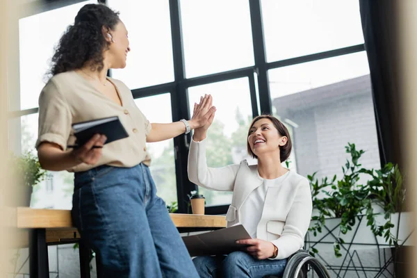 Happy businesswoman in wheelchair giving high five to african american colleague in office — Stock Photo