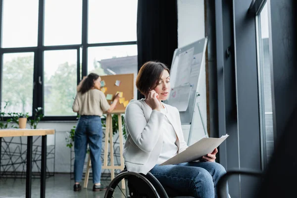 Businesswoman in wheelchair talking on mobile phone while blurred african american colleague working near corkboard — Stock Photo